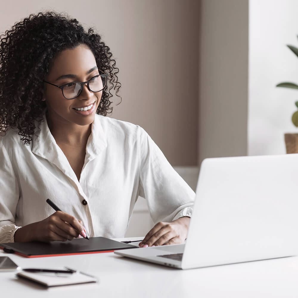 women who is writing notes on her tablets smiles while reading her laptop